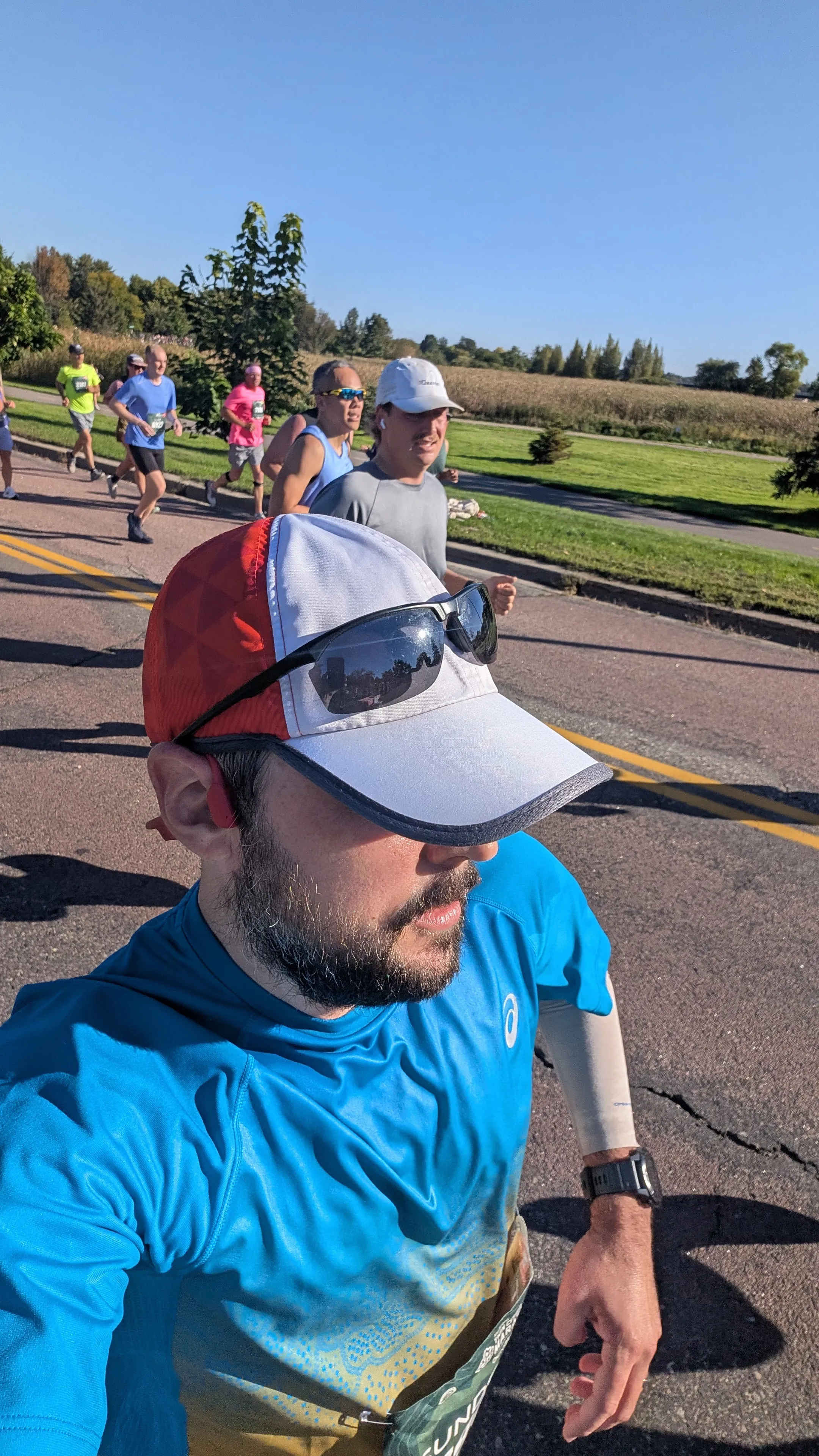 A selfie of me running the marathon in a blue shirt and red-and-white cap, with other runners and a cornfield behind me